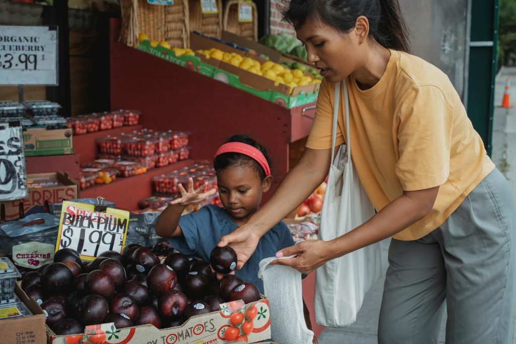 Asian woman putting black plum into eco bag while choosing fruits from box in street market with daughter