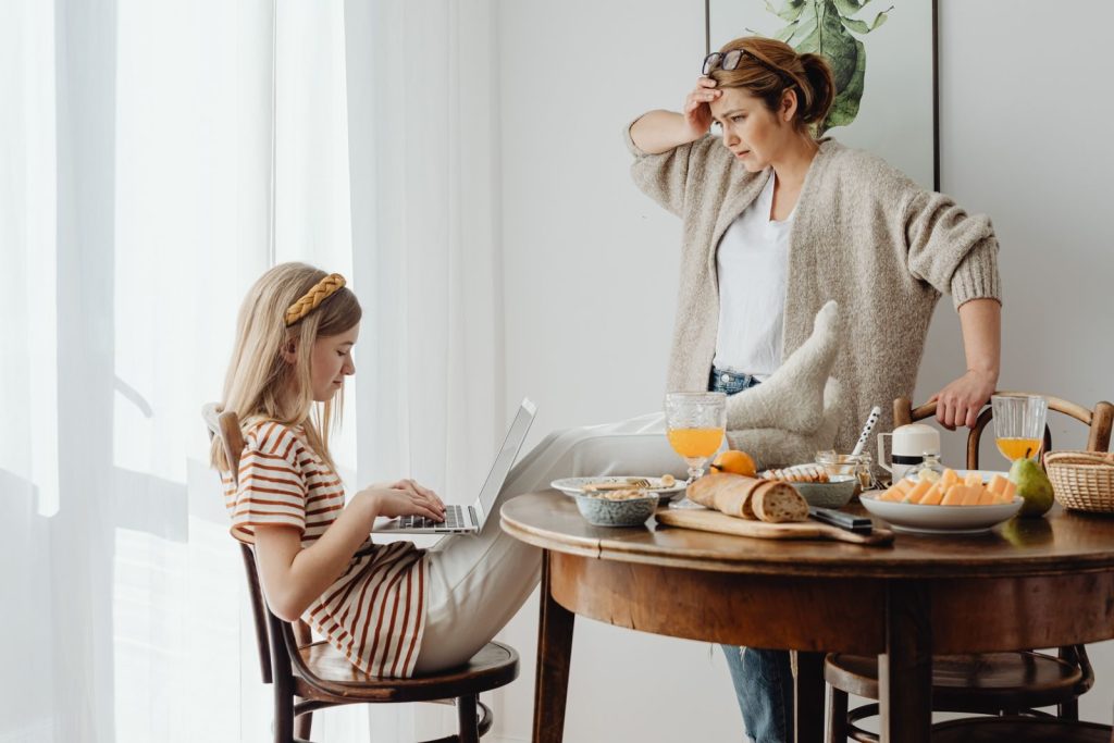 A mother and daughter engage in a morning routine with a laptop, breakfast, and an intimate moment indoors.