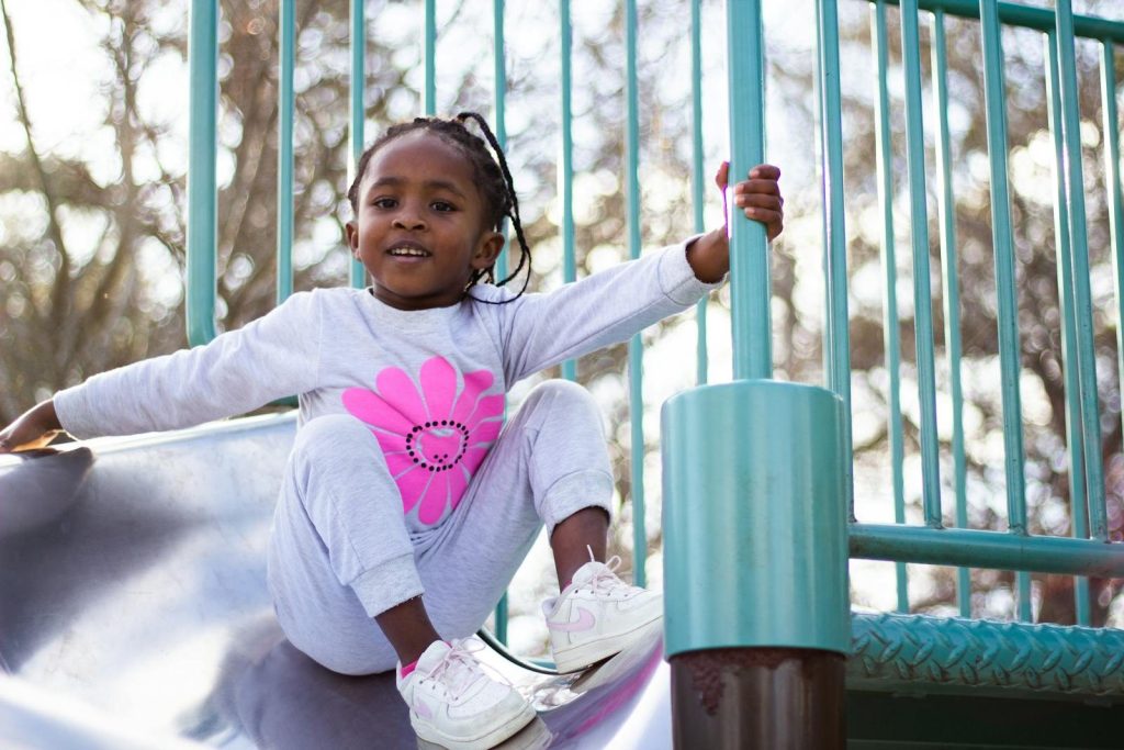 Young girl in a sweatsuit smiling while sliding down a playground slide on a sunny day.
