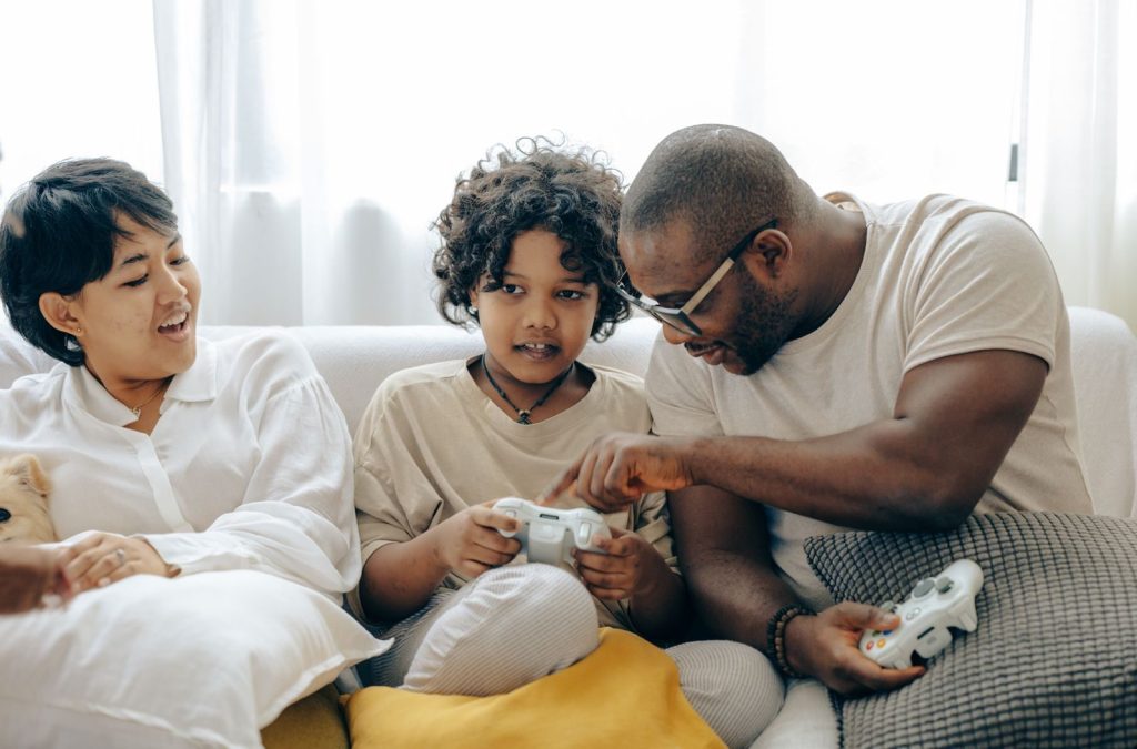Concentrated black father pointing on joystick buttons explaining rules of playing to son while positive mother watching process