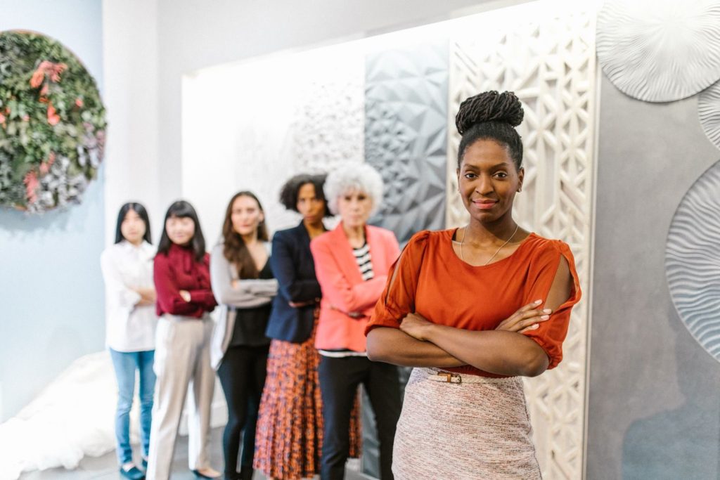 A confident female leader stands with diverse female colleagues in a modern office setting.