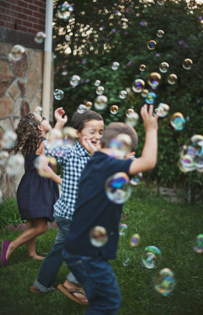 Three children playing with floating bubbles in a grassy backyard at golden hour