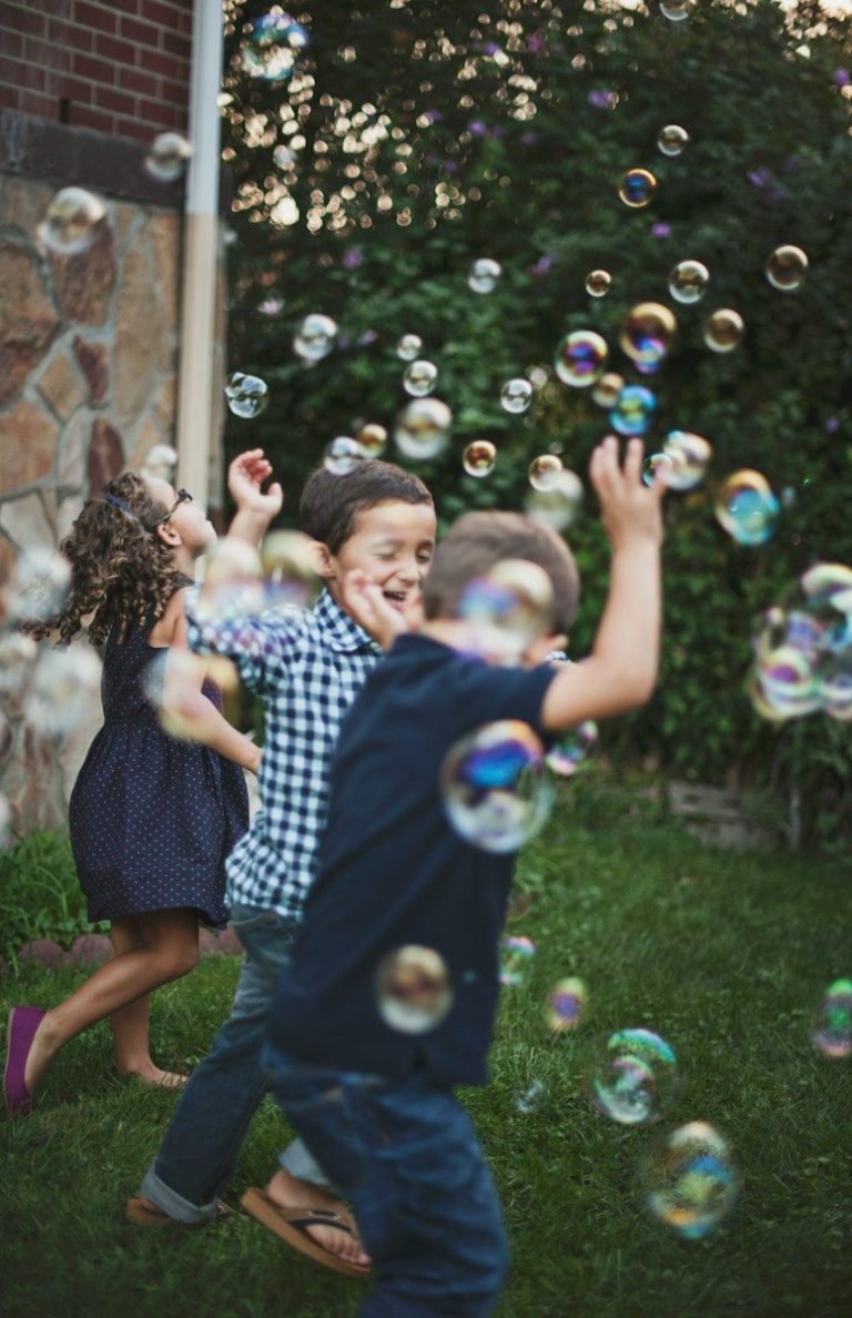 Three children playing with floating bubbles in a grassy backyard at golden hour